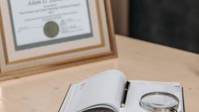 Open notebook with magnifying glass on wooden table beside framed certificate.