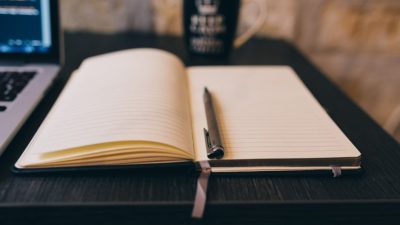 Open notebook with pen on a wooden desk, laptop, and a mug in a cozy setting.
