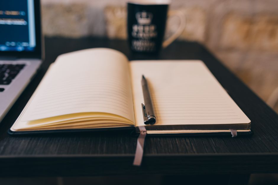 Open notebook with pen on a wooden desk, laptop, and a mug in a cozy setting.