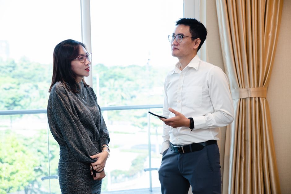 Two colleagues engaged in a business conversation indoors with a window view.