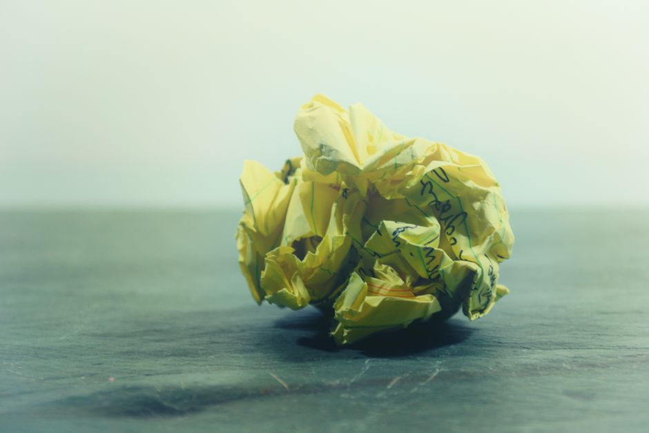 A close-up of a crumpled yellow paper ball with handwriting on a wooden surface.